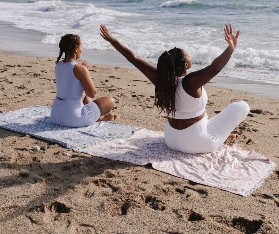 2 women at the beach with one hands raised and one sitting in a reflective mood.